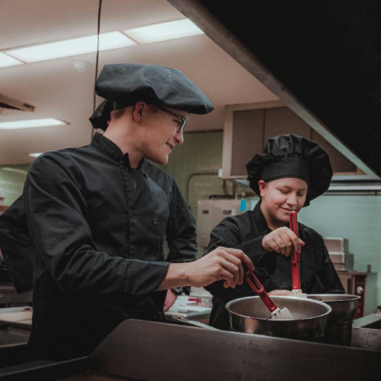 Dos chefs en una cocina profesional, uno con gafas y gorra, y otro con un gorro de chef, utilizando utensilios de cocina para preparar alimentos en ollas.
